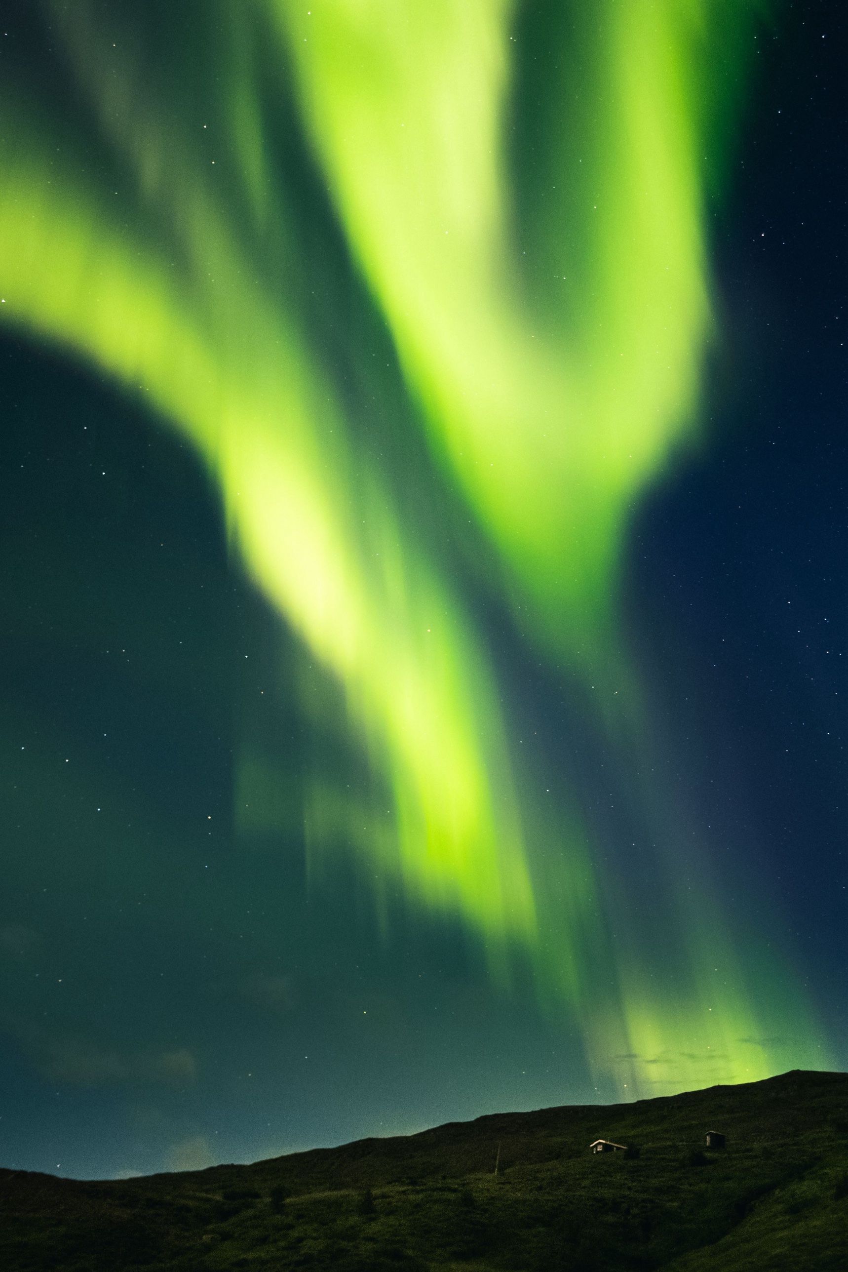 Bright green northern lights above a dark hillside with two small cabins