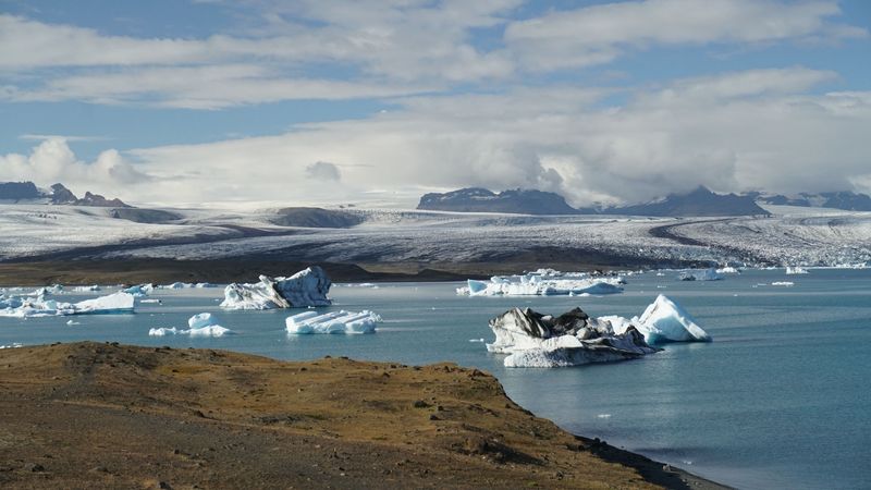 Icebergs in a turquoise lagoon below a wide glacier tongue and clouded mountains