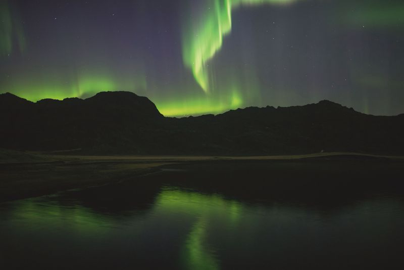 Green northern lights over dark mountains reflected in a lake