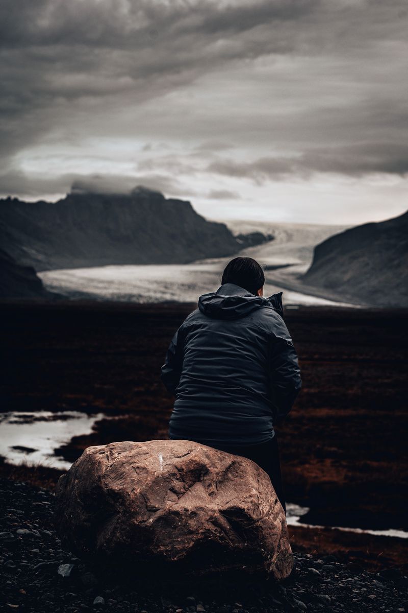 A walker sitting by a boulder looking out at a glacier tongue between dark mountains