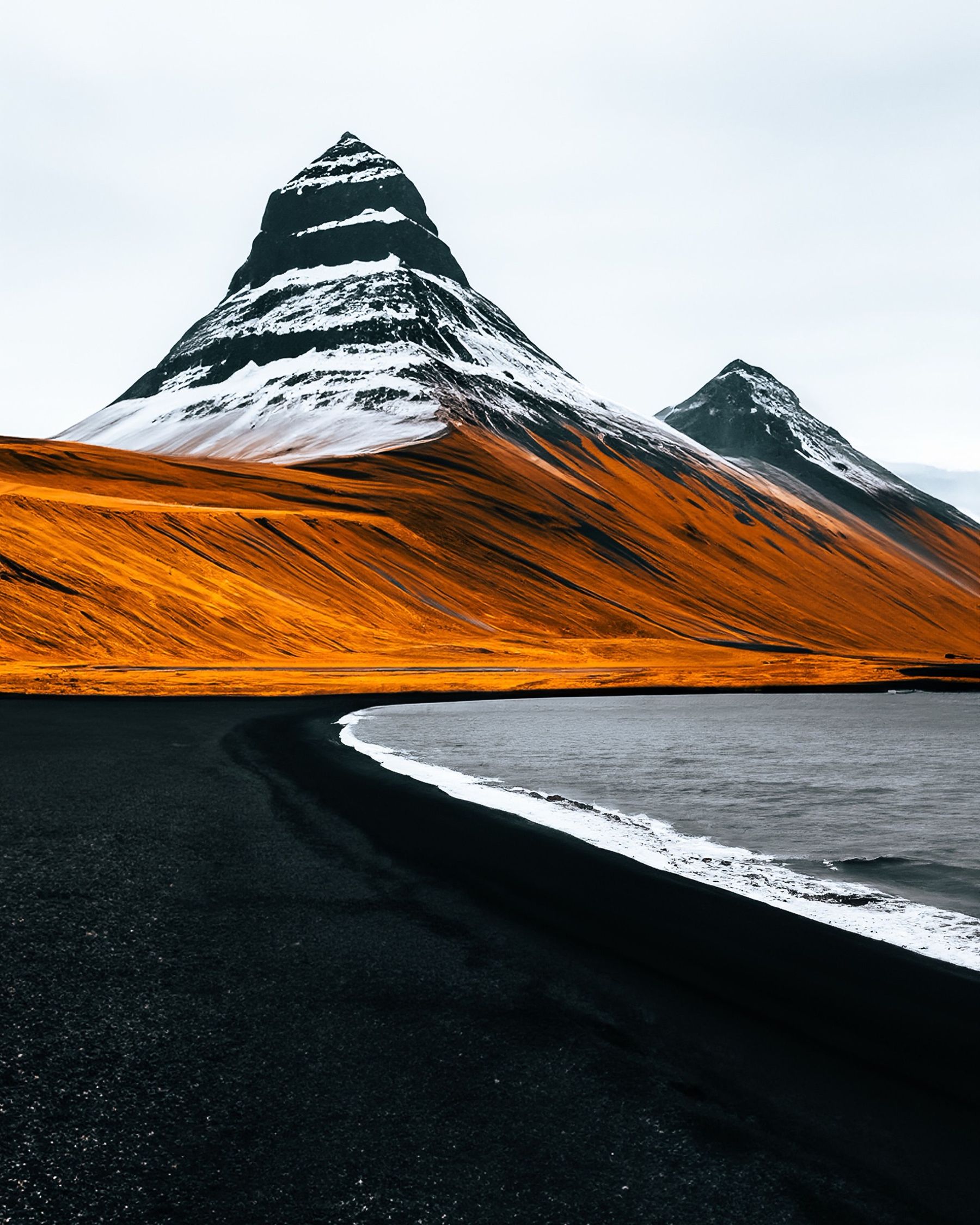 A snow-striped pyramid mountain above orange slopes and a curving black beach