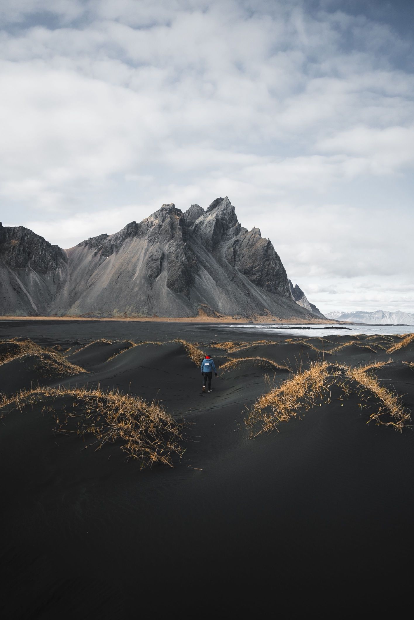A walker in a blue jacket crossing black dunes below the jagged peaks of Vestrahorn