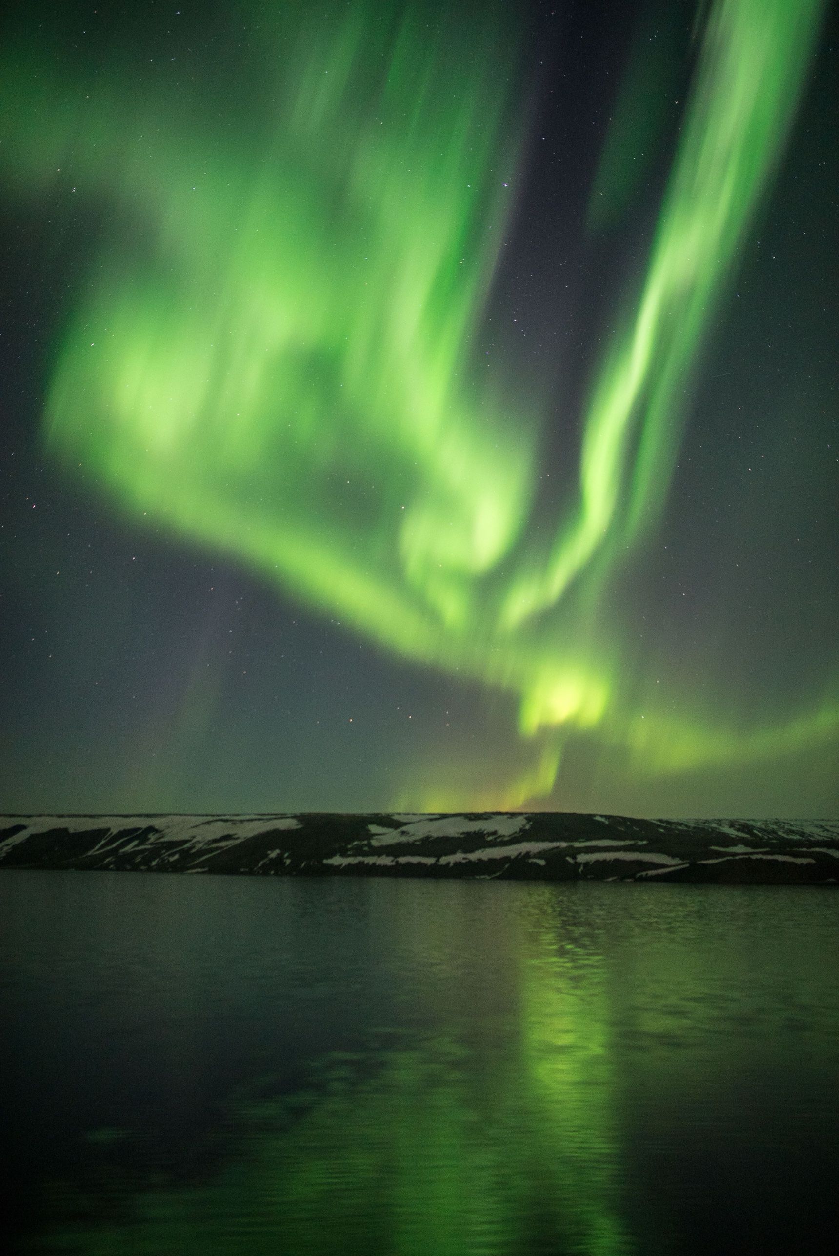 Green northern lights swirling over a snowy ridge, reflected in a calm fjord